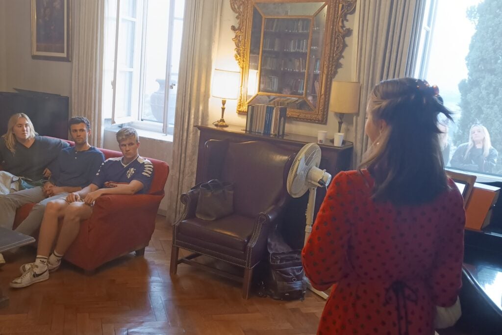 Three students sitting on a couch in the Villa's "Music Room" with a woman in a red dress standing at a pianoforte with her back to the camera.