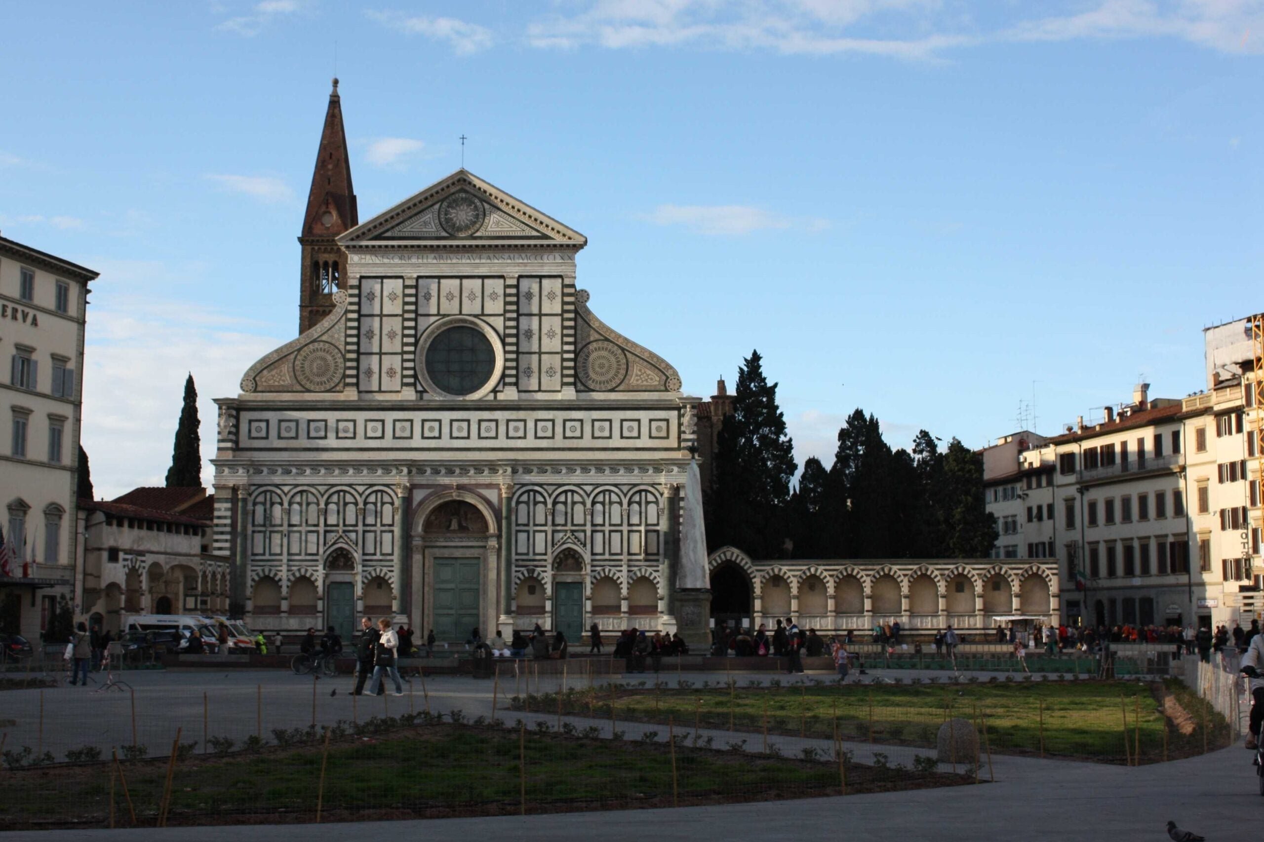 Renaissance basilica of Santa Maria Novella bathed in sunlight with pedestrians walking in front.