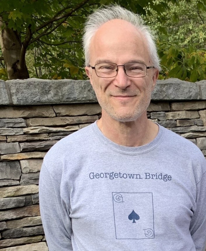 Prof. Astarita in a grey Georgetown Bridge shirt standing in front of a stone wall.