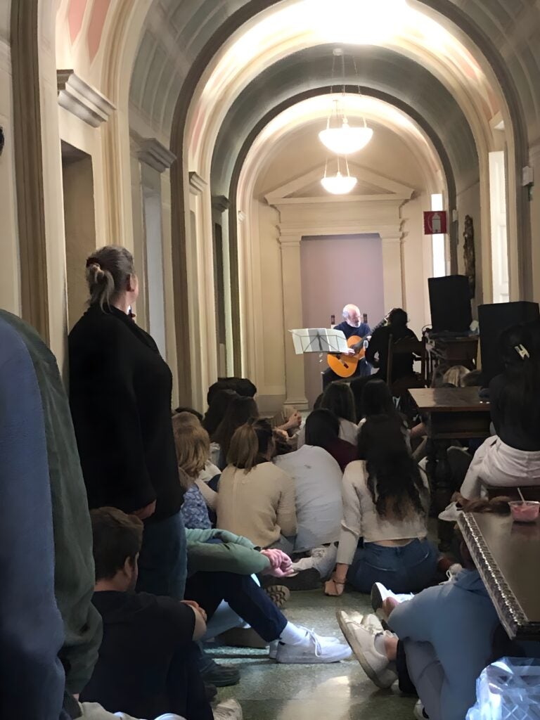 Students standing and sitting on the floor in a hallway with a man playing guitar in the background.